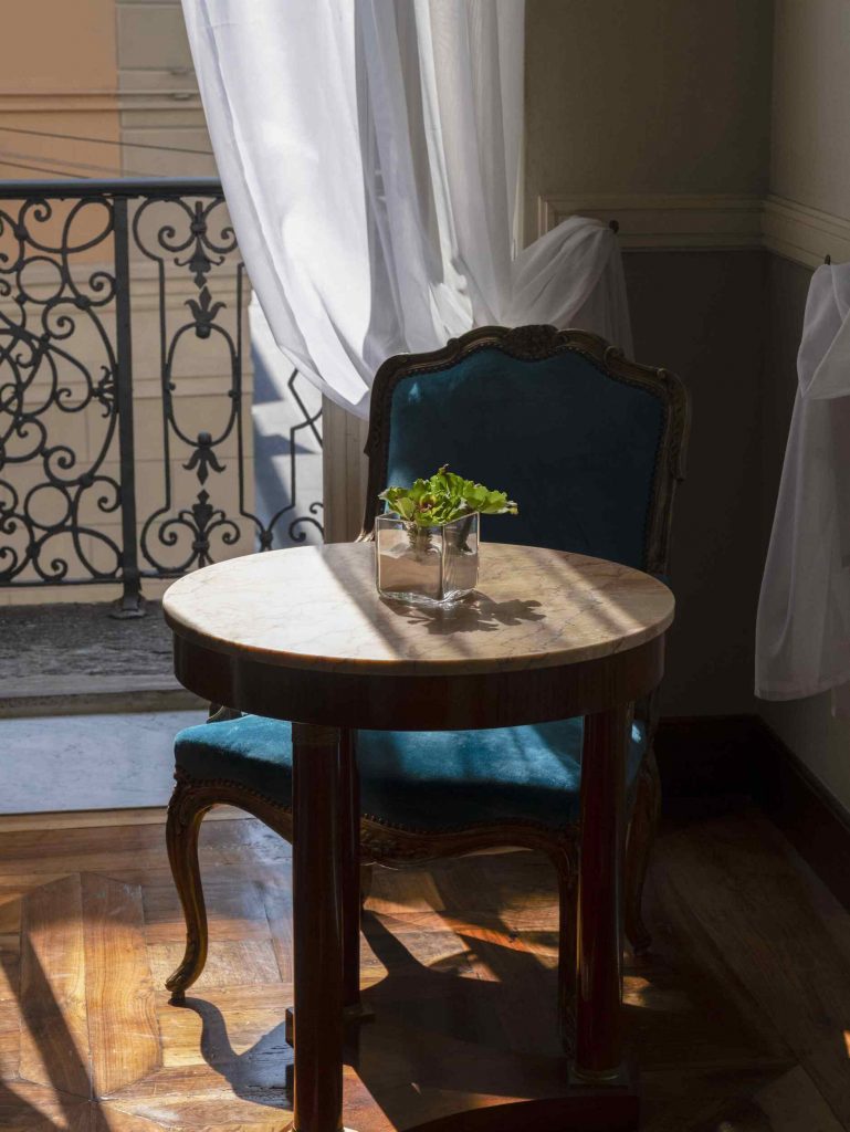 vue suggestive du salon de la suite avec table basse et fauteuil tapissé de velours turquoise foncé. La prise de vue est à contre-jour, la lumière du soleil filtrant à travers la porte-fenêtre à l'arrière-plan, aux rideaux blancs ouverts, et éclairant la table basse sur laquelle est posé un petit pot contenant une plante succulente