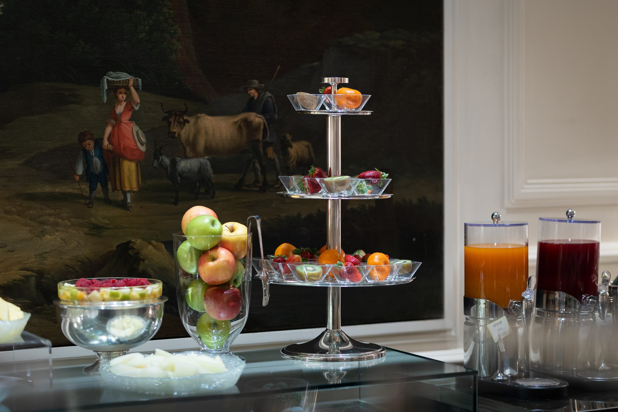 buffet avec fruits frais mélangés sur un support en cristal, vase avec pommes rouges, jaunes et vertes, salade de fruits servie dans une coupe en cristal et argent.
